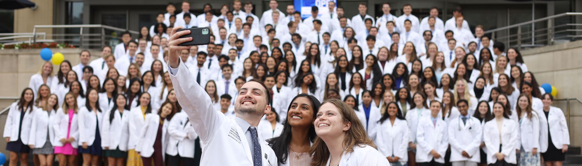Three students take selfies with a group of students on the steps of Gelman Library